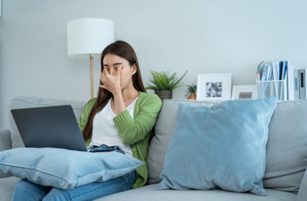 A woman sitting on her couch with her laptop on her lap, and she is also rubbing her eyes with her hand due to eye strain
