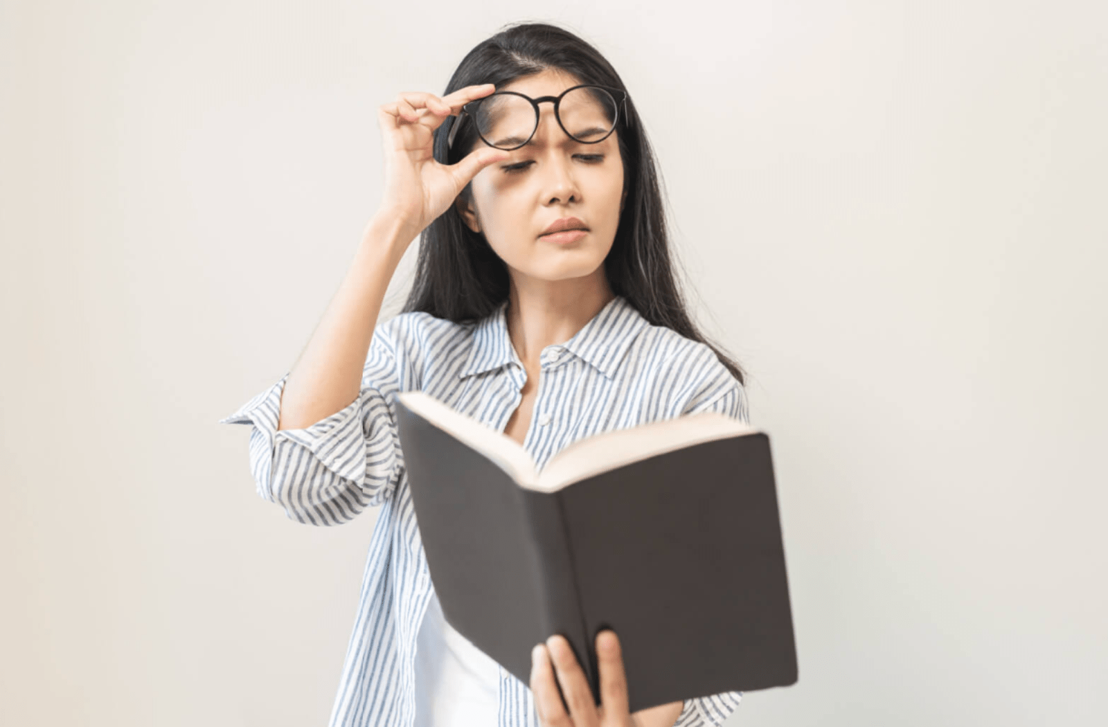 A woman trying to read a book, and she's holding up her glasses because it's hard to see the book