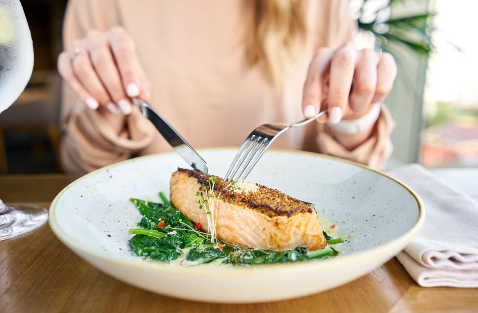 A close-up of a salmon steak fillet being cut by a woman using a fork and knife.