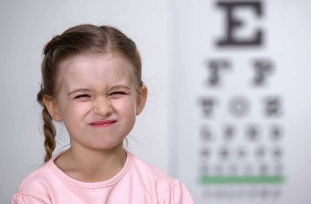 A young girl sitting in front of a Snellen eye chart and she is squinting due to vision issues