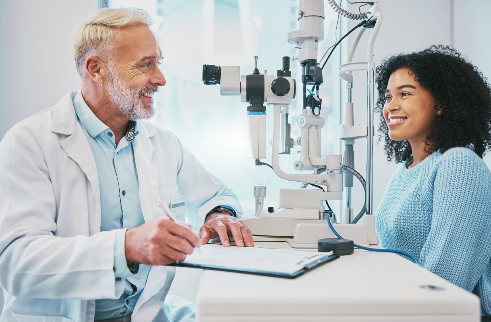 A male optometrist and a young female patient smiling while discussing the results of her eye exam.