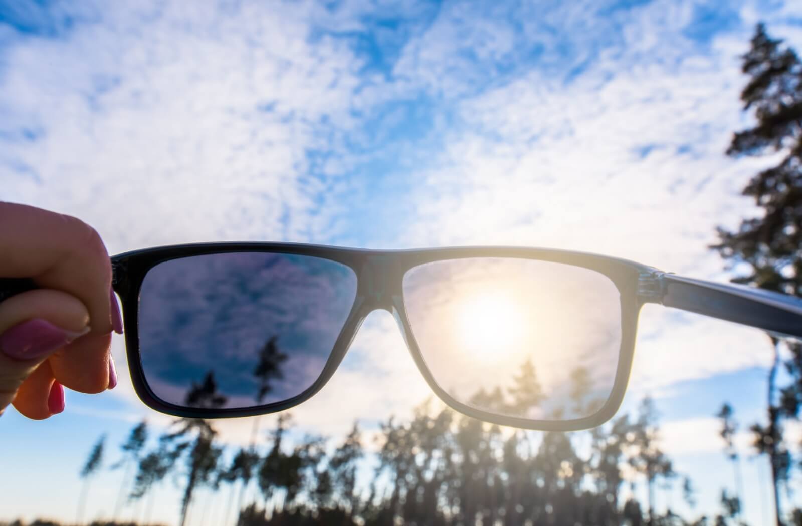 A pair of sunglasses that is being held up blocks the sun from a sunny sky with a grove of trees in the background