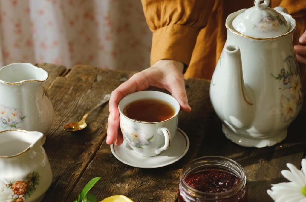 An individual places a full cup of tea onto a wooden table alongside a flowery tea set and an open jar of jam