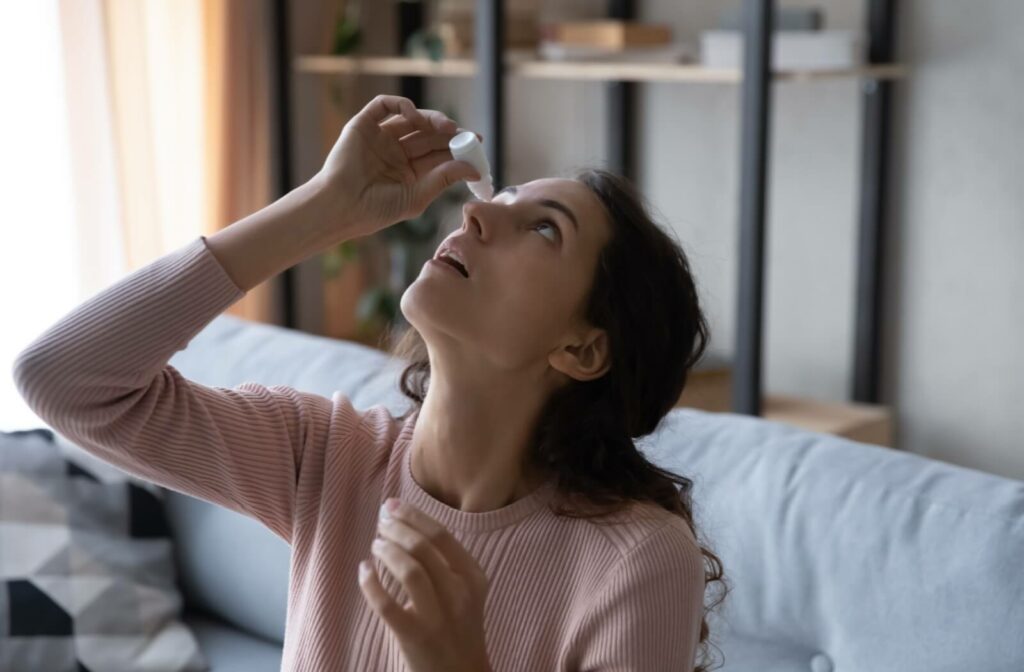 A person sitting on their couch applying eye drops to their right eye to help remove a stuck inside-out contact lens.
