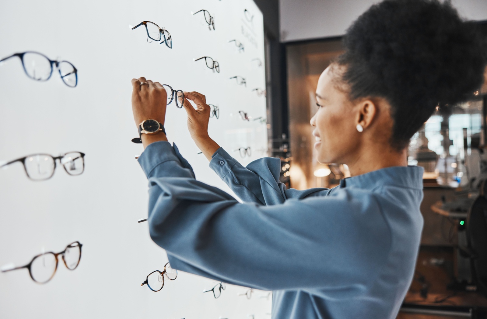 Person selecting eyeglass frames from a display wall in an optical store.