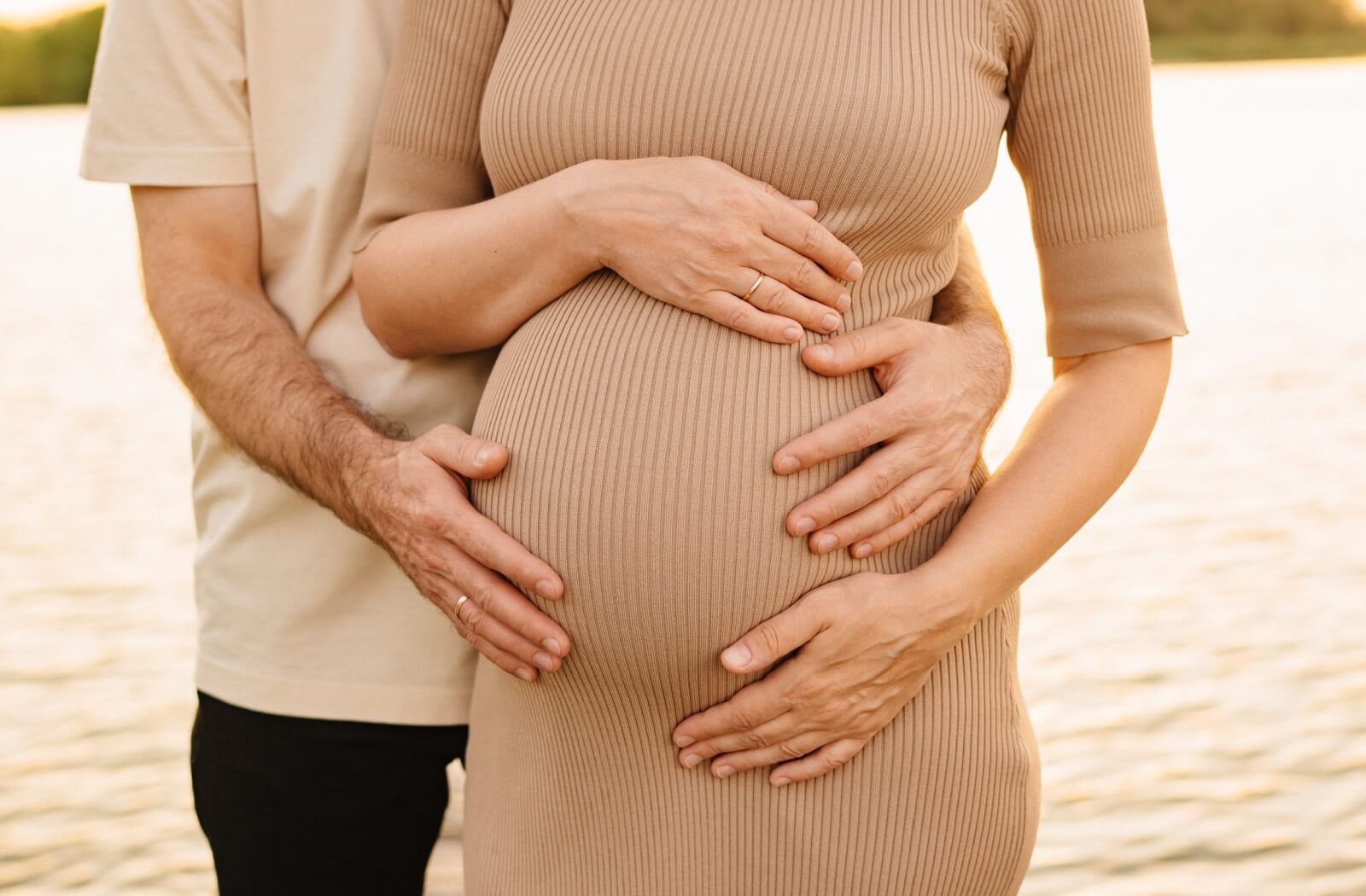 Close-up of a pregnant woman holding her belly with her partner’s hands gently placed over hers.