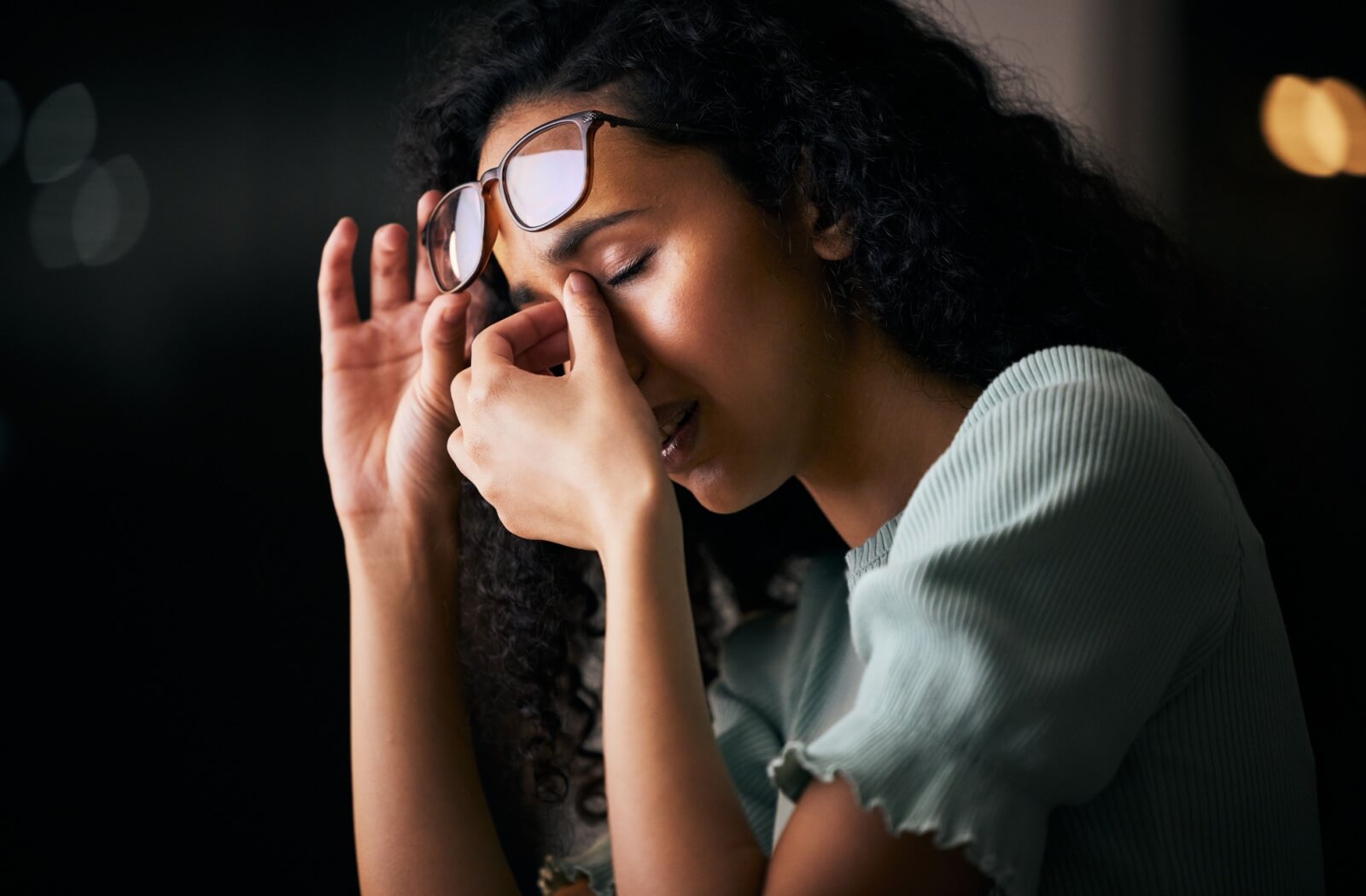 A person with curly hair lifts their glasses while rubbing the bridge of their nose, appearing to experience eye discomfort in a dimly lit setting.