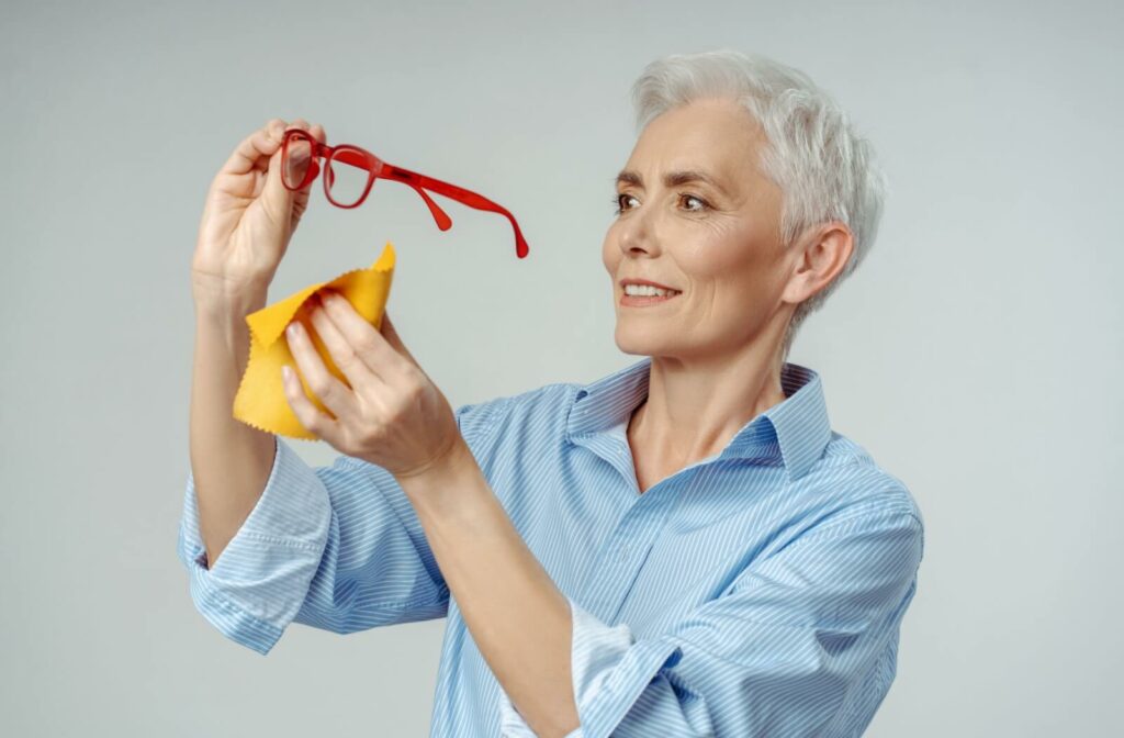 A person with short gray hair holding red eyeglasses and cleaning the lenses with a yellow cloth while smiling.