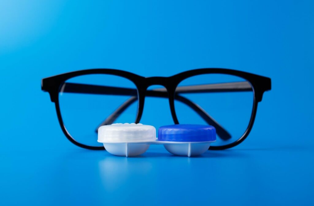 Black eyeglasses placed behind a contact lens case with blue and white lids on a solid blue background, representing contact lenses versus glasses for vision correction.