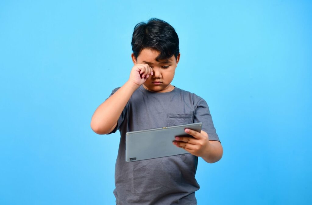 A child wearing a gray shirt rubbing one eye while holding and looking at a tablet against a solid blue background.