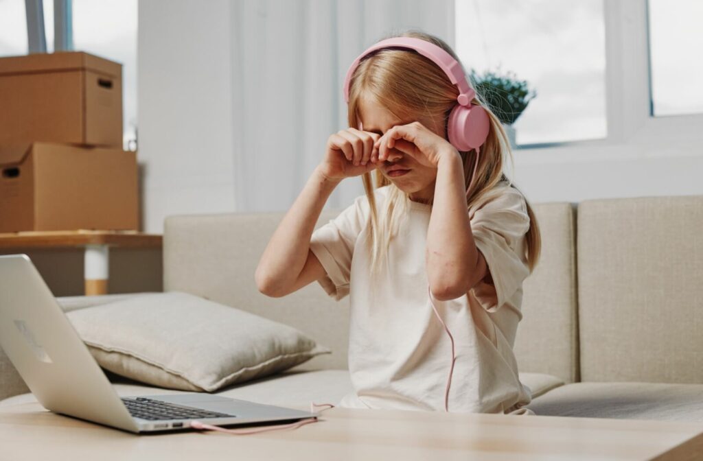 A young girl wearing pink headphones sitting in front of a laptop and rubbing her eyes in discomfort.