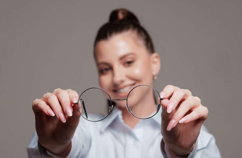 A woman holding a pair of thin, round wire-frame eyeglasses toward the camera, demonstrating frame shapes for face shape consultations.