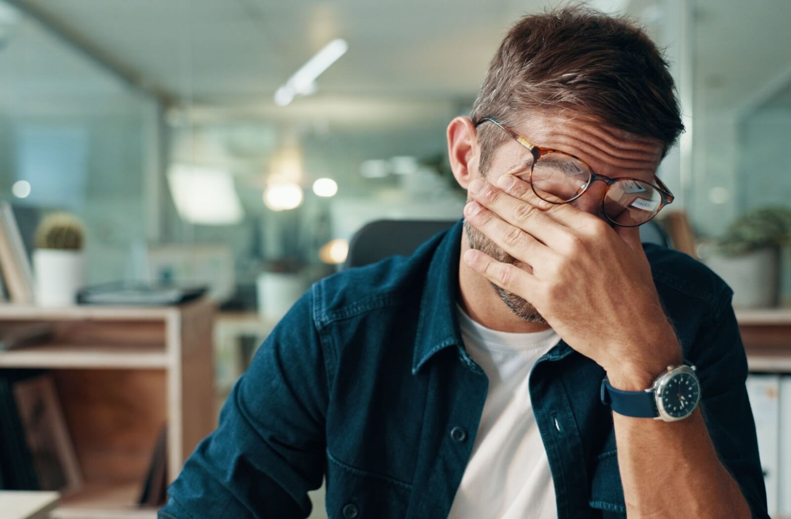 A man wearing eyeglasses rubbing his tired eyes while sitting at an office desk, illustrating chronic eye fatigue and digital strain.