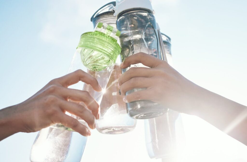 Several hands holding up reusable water bottles in a celebratory toast against a bright sky, illustrating the importance of hydration for eye health and energy.
